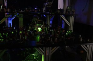 A photo of the optical components used to trap and manipulate atoms in the JILA neutral-atom qubit experiment. The photo is dark and bathed in green and blue light from the lasers used in the experiment.