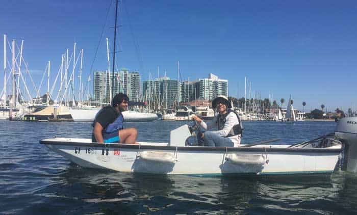 Photo of graduate students Shahank Gowda and Muchen Xu sitting in a small boat with an outboard motor, in a harbour with palm trees and the masts of sailing vessels visible in the background